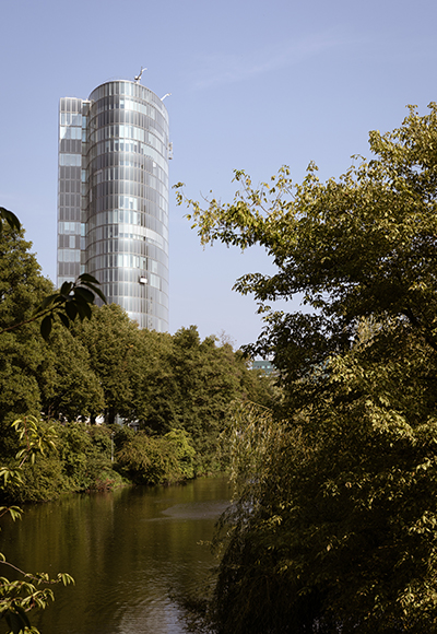 Moderne Architektur – Glas-Wolkenkratzer in naturnaher Umgebung Ein moderner Glas-Wolkenkratzer, umgeben von Bäumen und einem Fluss, aufgenommen an einem sonnigen Tag. Das Bild kombiniert Natur und moderne Architektur.