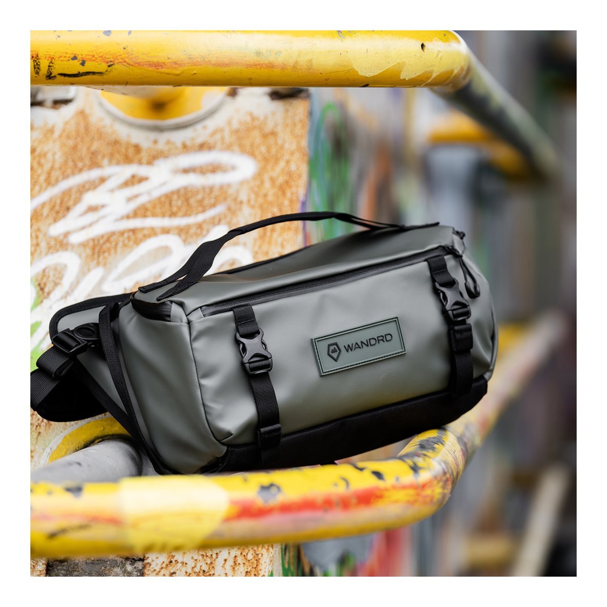 WANDRD ROGUE Sling 9L Wasatch Green - Vorderansicht: Graue WANDRD Sling Bag mit Logo und Schnallen auf rostigem gelben Geländer.
