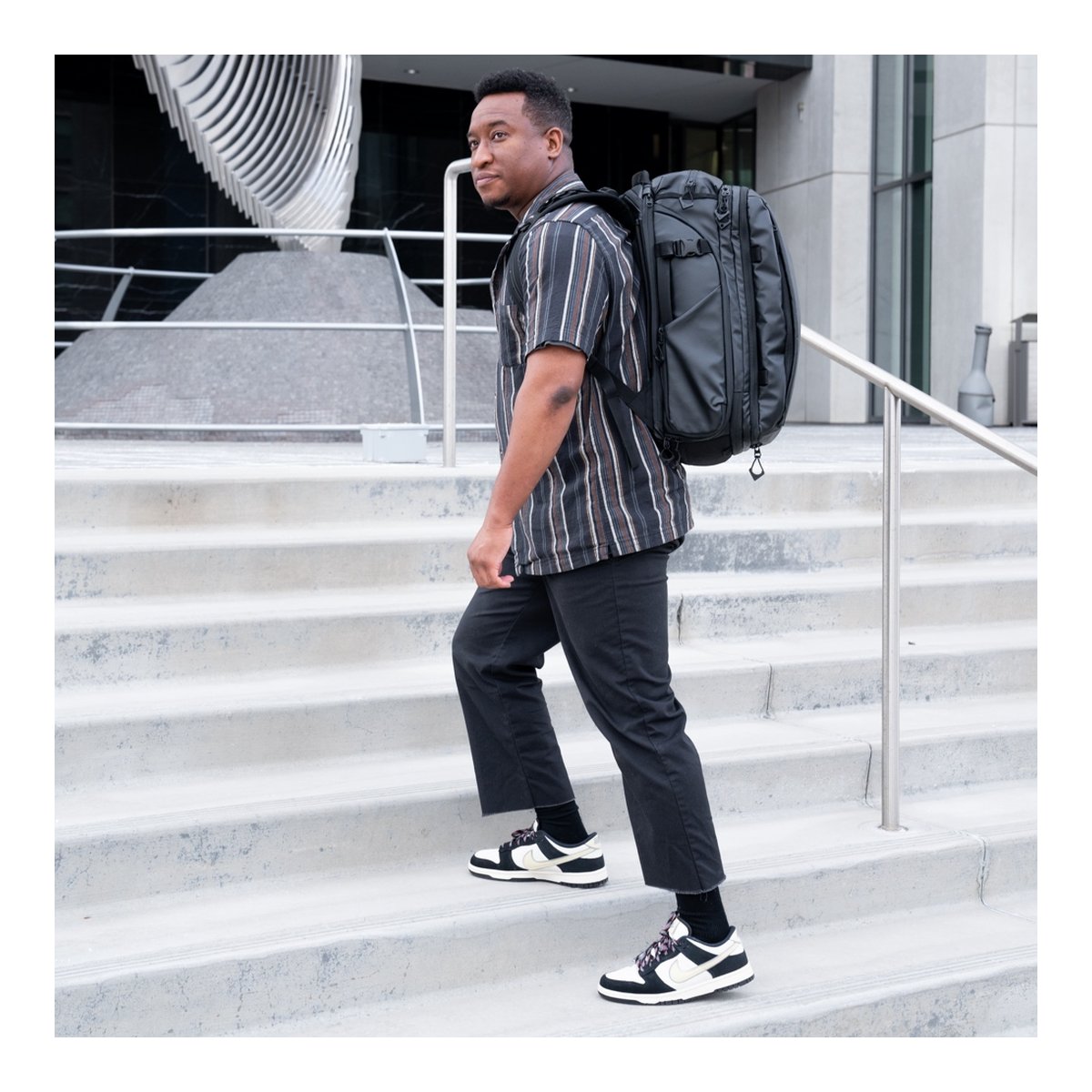 WANDRD Transit 35L Travel Backpack Wasatch Green Essential Bundle - Anwendungsbeispiel: Mann trägt schwarzen Rucksack auf dem Rücken beim Treppensteigen. WANDRD Transit 35L Travel Backpack Wasatch Green Essential Bundle - Anwendungsbeispiel: Mann trägt schwarzen Rucksack auf dem Rücken beim Treppensteigen.