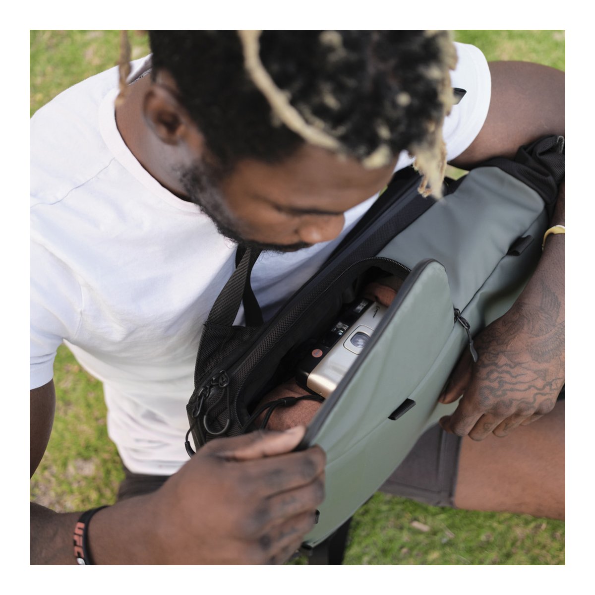 WANDRD PRVKE Lite 11-Liter Green - Anwendungsbeispiel: Mann öffnet graue Sling Bag mit Kamera und Trinkflasche WANDRD PRVKE Lite 11-Liter Green - Anwendungsbeispiel: Mann öffnet graue Sling Bag mit Kamera und Trinkflasche