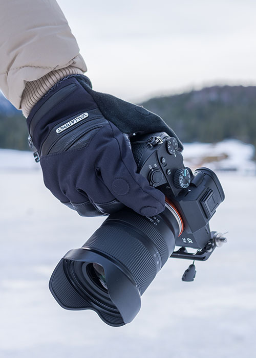 Hand mit Winter-Fotohandschuh hält eine Kamera im verschneiten Außenbereich, Fokus auf sicherem Griff und Bedienung.