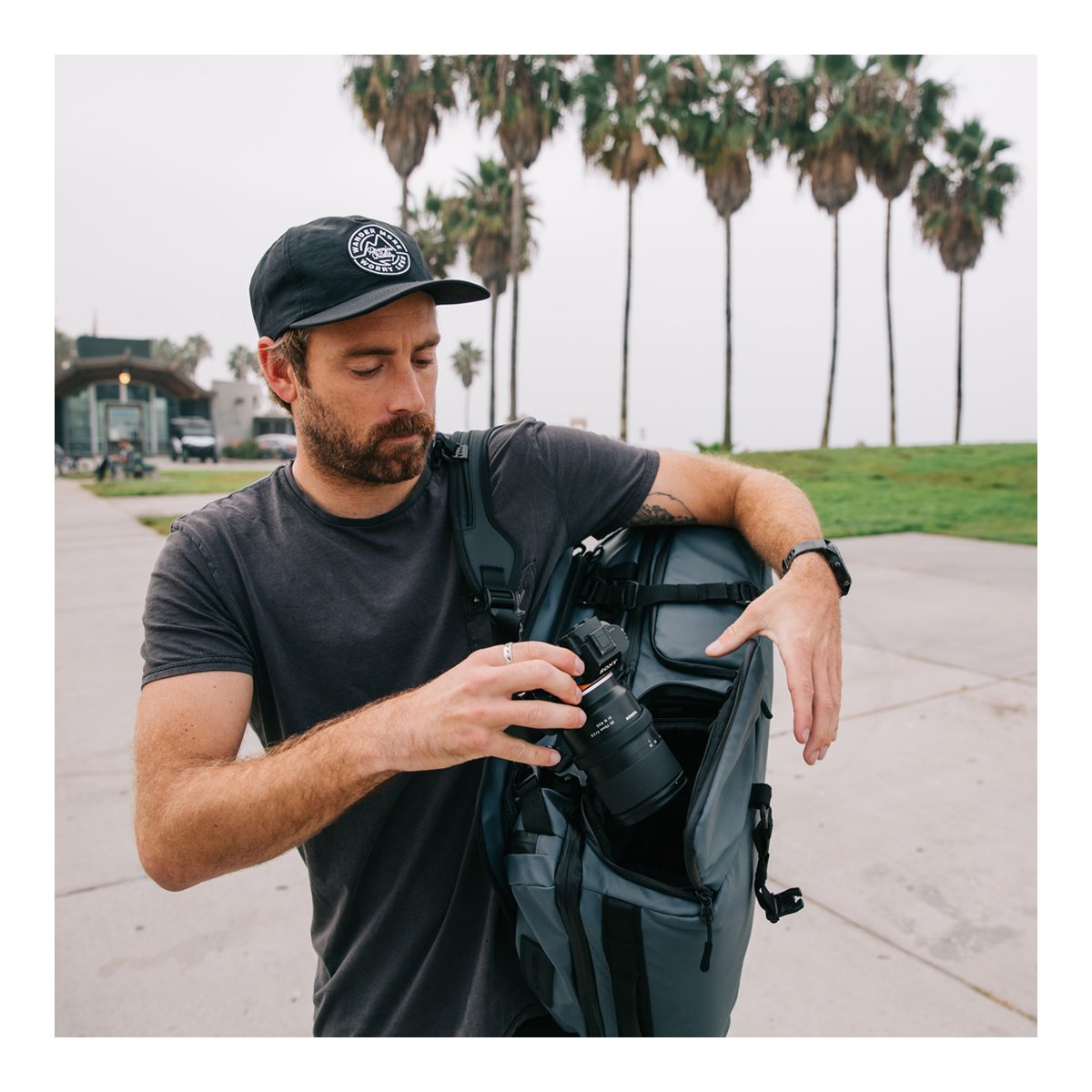 WANDRD HEXAD ACCESS DUFFEL Aegean Blue - Anwendungsbeispiel: Mann entnimmt Kamera aus grauem Rucksack. Palmen im Hintergrund.