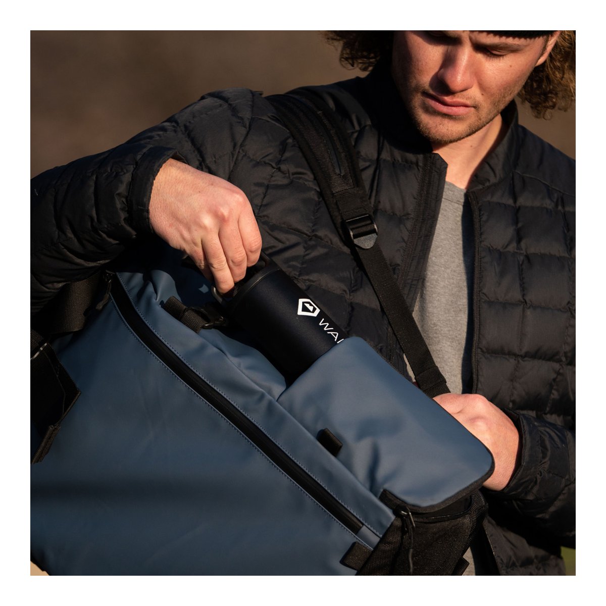 WANDRD PRVKE 21-Liter Blue V3 - Anwendungsbeispiel: Person verstaut schwarze Trinkflasche mit WAI-Logo in blauer Tasche.
