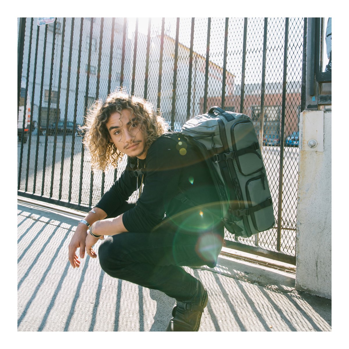 WANDRD HEXAD ACCESS DUFFEL Wasatch Green - Anwendungsbeispiel: Mann trägt grauen Rucksack und hockt vor Gitterzaun im Sonnenlicht WANDRD HEXAD ACCESS DUFFEL Wasatch Green - Anwendungsbeispiel: Mann trägt grauen Rucksack und hockt vor Gitterzaun im Sonnenlicht