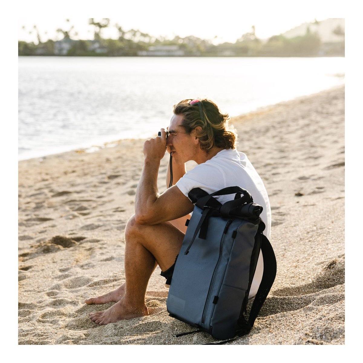 WANDRD PRVKE Lite 11-Liter Blue - Anwendungsbeispiel: Grauer Roll-Top-Rucksack am Strand neben einer Person mit Fernglas. WANDRD PRVKE Lite 11-Liter Blue - Anwendungsbeispiel: Grauer Roll-Top-Rucksack am Strand neben einer Person mit Fernglas.