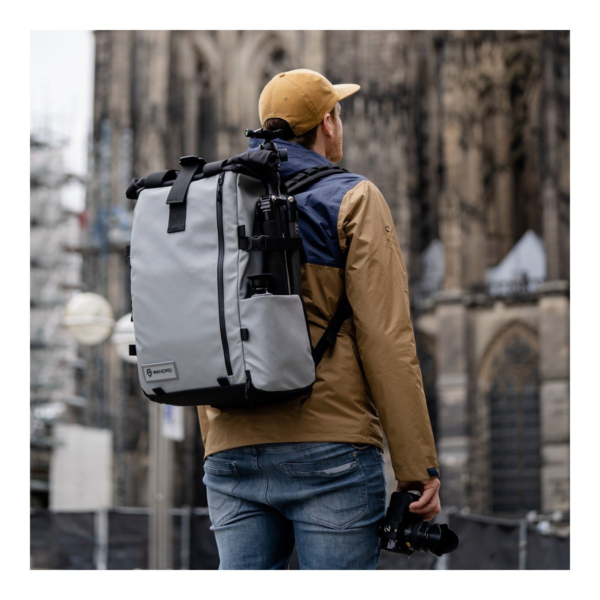 WANDRD PRVKE 41-Liter DisNet Grey V3 - Anwendungsbeispiel: Person mit grauem Fotorucksack und befestigtem Stativ, Kamera in der Hand.