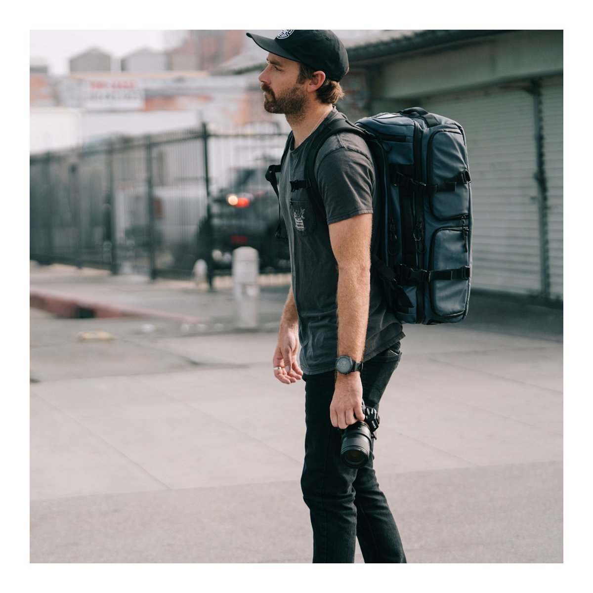 WANDRD HEXAD ACCESS DUFFEL Aegean Blue - Anwendungsbeispiel: Mann trägt blauen Rucksack auf dem Rücken, Seitenansicht, hält Kamera.