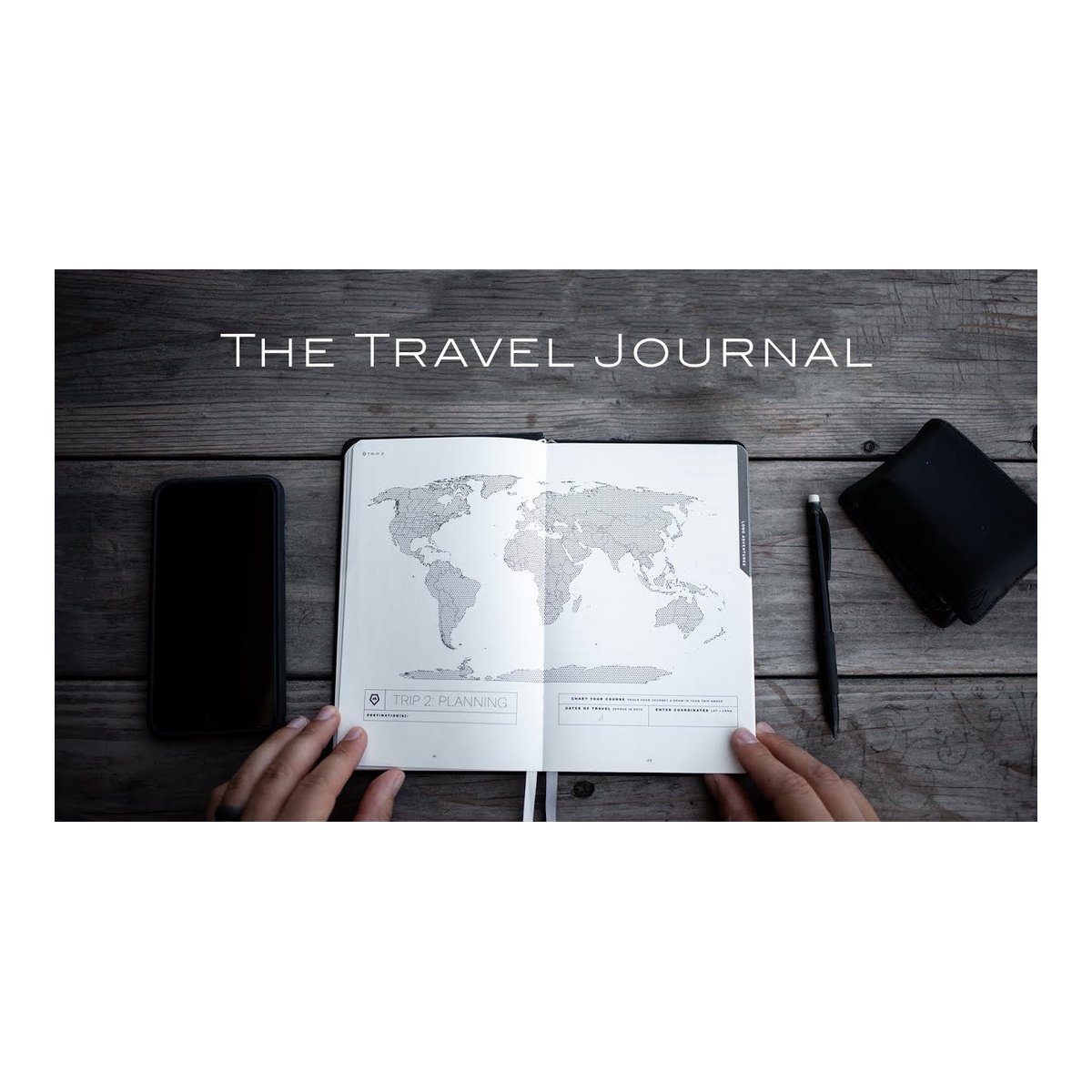WANDRD THE TRAVEL JOURNAL Black - Anwendungsbeispiel: Geöffnetes Travel Journal mit Weltkarte und Händen beim Reiseplanen auf Holztisch, dazu Handy, Stift, Geldbörse.