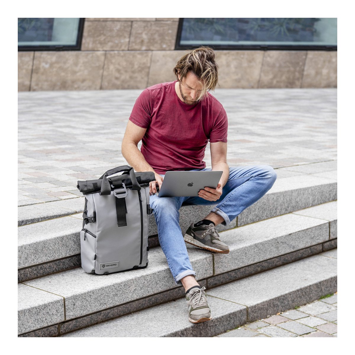 WANDRD PRVKE 31-Liter DisNet Grey Photo Bundle - Anwendungsbeispiel: Mann sitzt mit Laptop neben grauem Roll-Top-Rucksack auf Steinstufen.