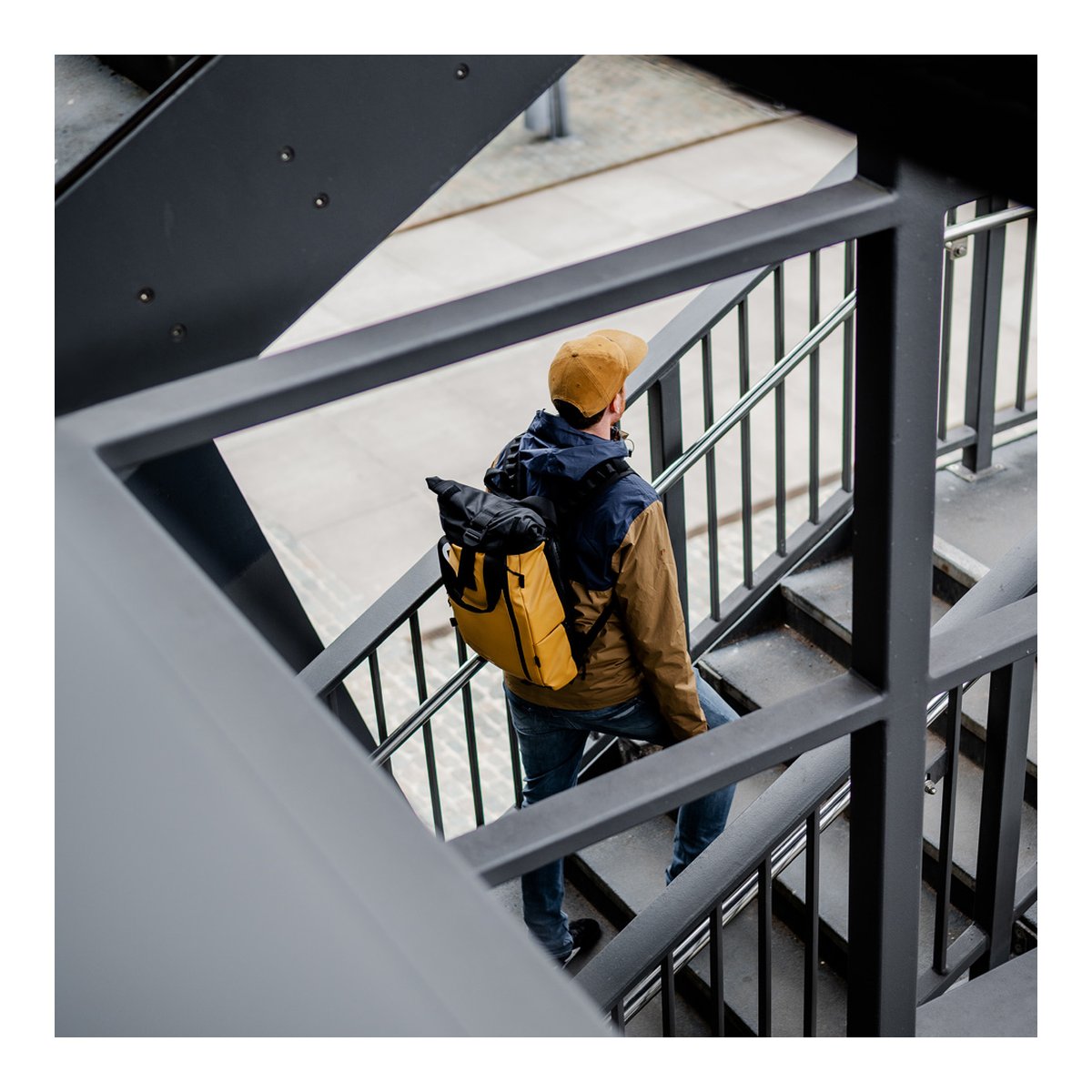 WANDRD PRVKE Lite 11-Liter Dallol Yellow - Anwendungsbeispiel am Rücken: Mann trägt gelb-schwarzen Roll-Top-Rucksack beim Treppensteigen.