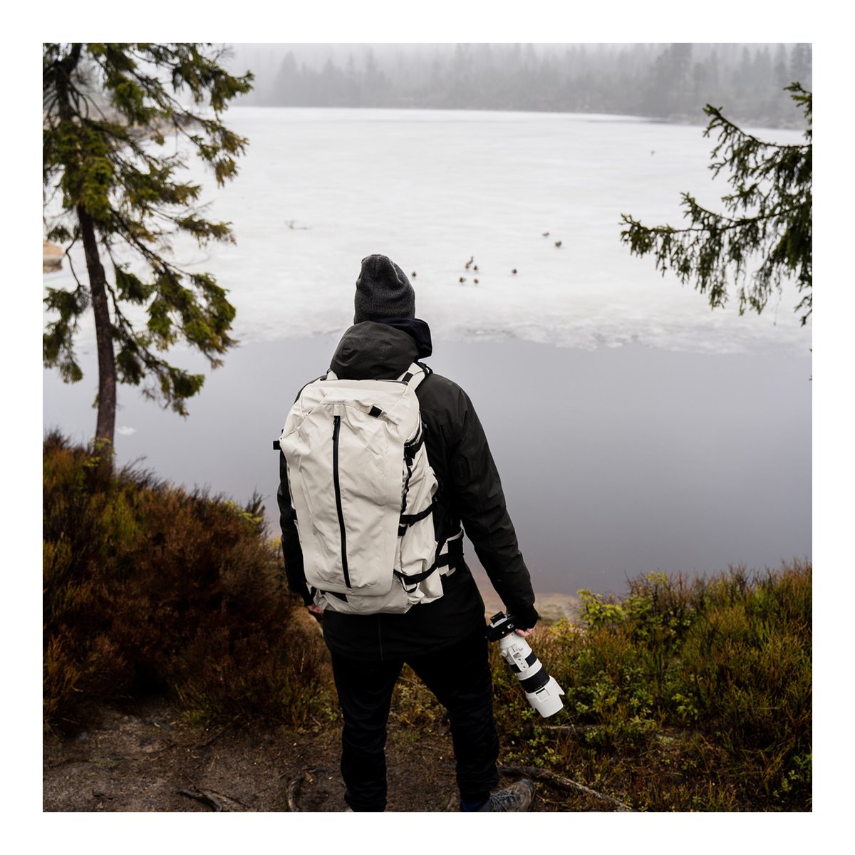 Wandrd Fernweh M/L Tan Photo Bundle - Anwendungsbeispiel Rückansicht: Person mit beigem Rucksack und Kamera vor winterlicher See-Landschaft.