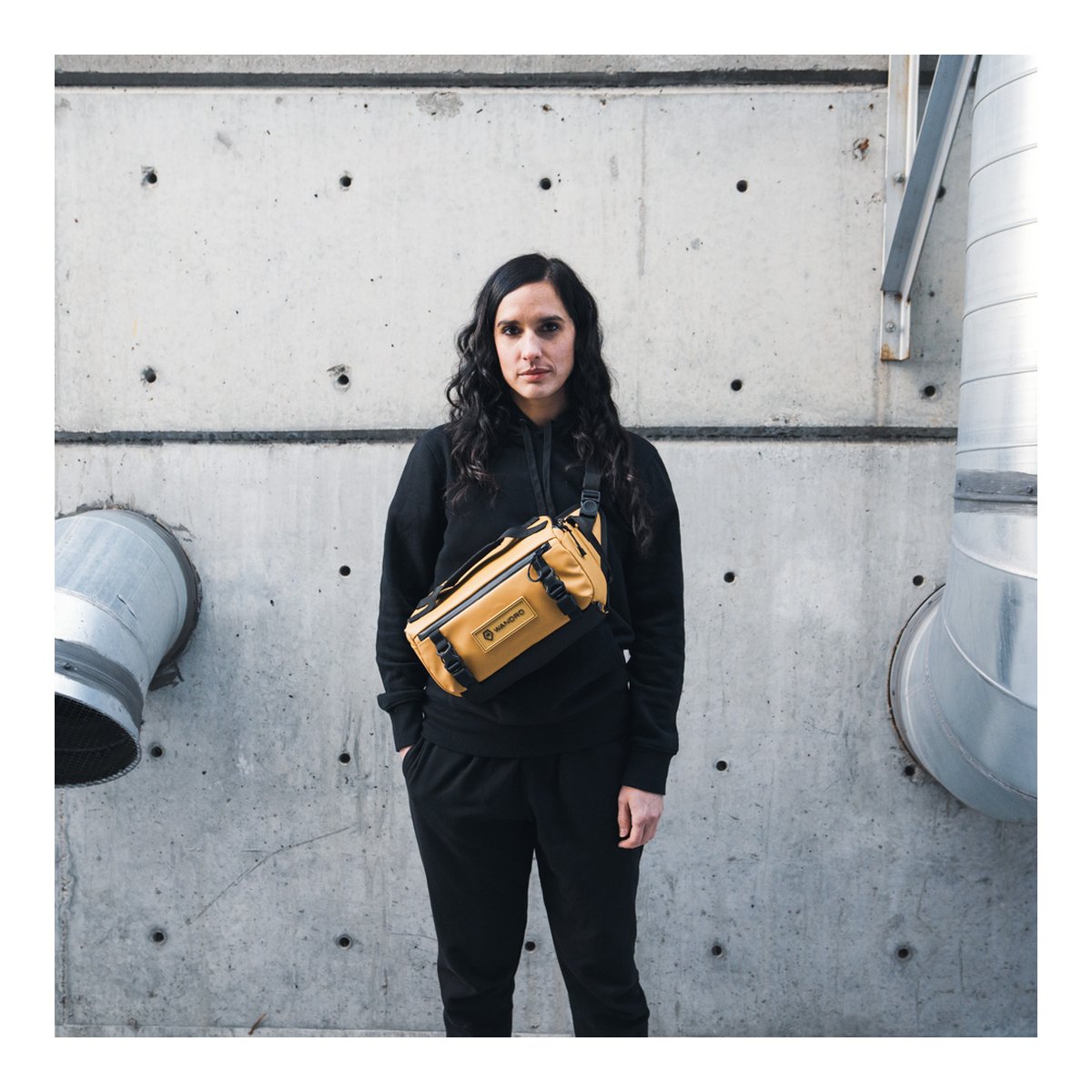 WANDRD ROGUE Sling 9L Dallol Yellow - Anwendungsbeispiel: Frau trägt senffarbene Sling Bag quer über der Brust vor Betonwand. WANDRD ROGUE Sling 9L Dallol Yellow - Anwendungsbeispiel: Frau trägt senffarbene Sling Bag quer über der Brust vor Betonwand.