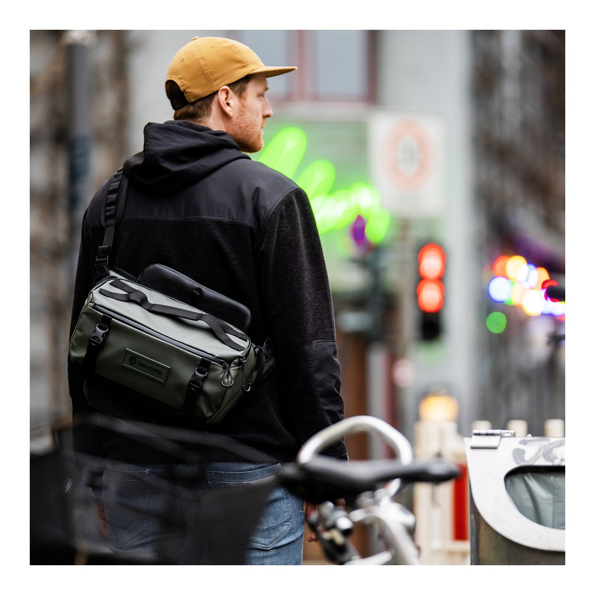 WANDRD ROGUE Sling 9L Wasatch Green - Anwendungsbeispiel: Grüne Sling Bag an Mann, von hinten/seitlich in städtischer Umgebung getragen.