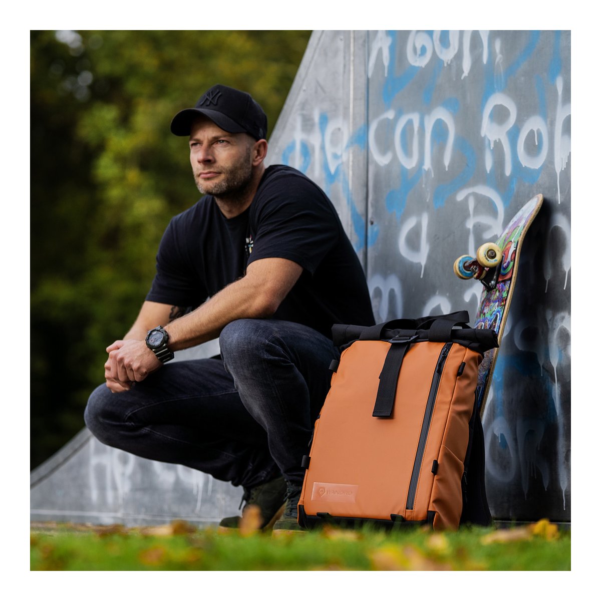 WANDRD PRVKE Lite 11-Liter Sedona Orange - Anwendungsbeispiel: Mann hockt neben orangefarbenem Rucksack und Skateboard an einer Betonwand.