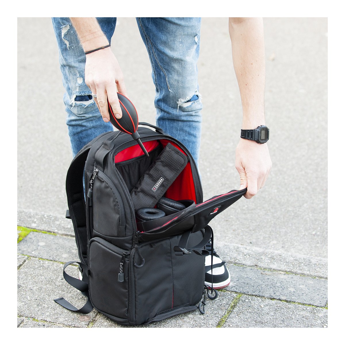 Caruba Skydex 10 - Anwendungsbeispiel: Person verstaut Kamera-Blasebalg und Kopfhörer im geöffneten Fotorucksack mit rotem Innenfutter.