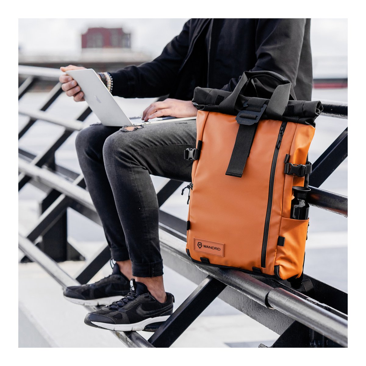 WANDRD PRVKE 21-Liter Sedona Orange Photo Bundle V3 - Anwendungsbeispiel: Orangefarbener Roll-Top Rucksack neben einer Person mit Laptop auf Geländer.