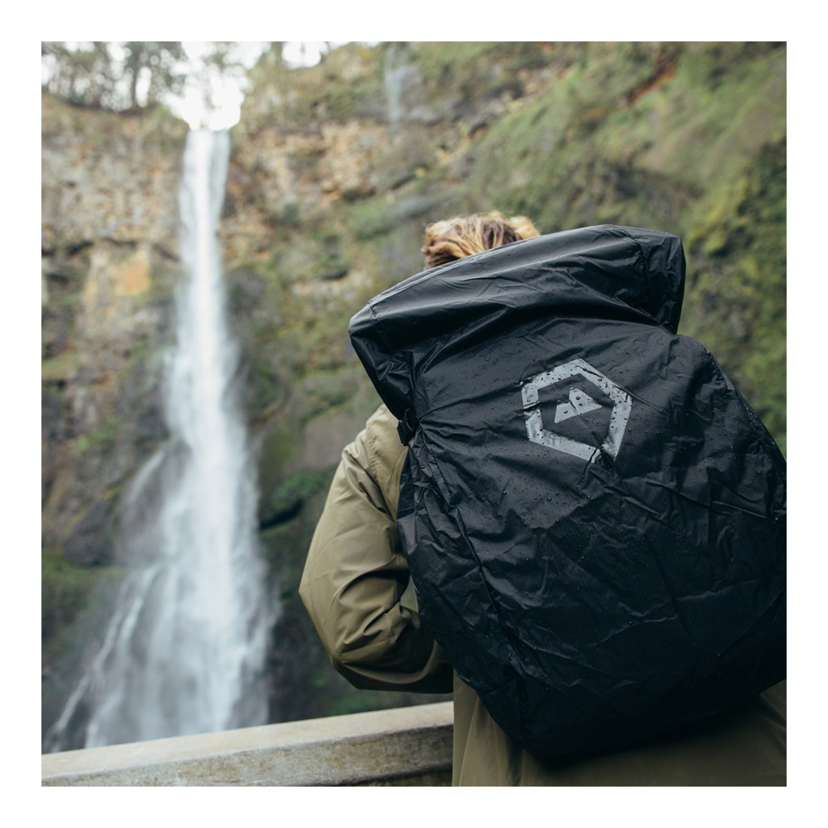WANDRD RAINFLY - Anwendungsbeispiel: Rückansicht wasserdichter Rucksack mit Logo und Wassertropfen vor Wasserfall.