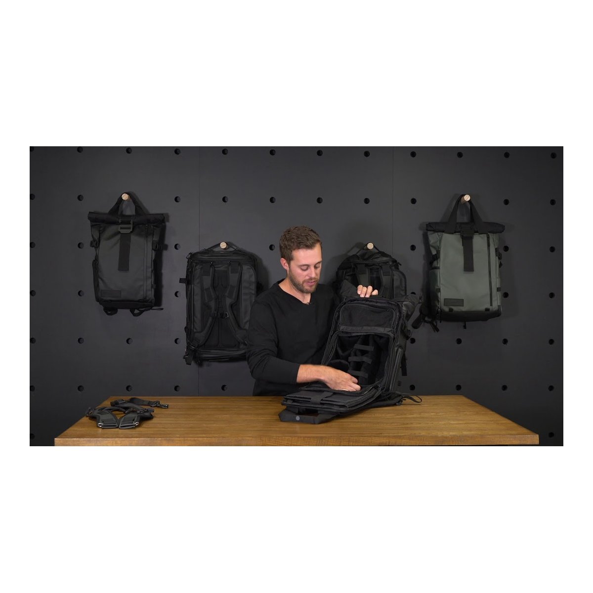 WANDRD Camera Cube Pro+ (31 Liter PRVKE) - Mann demonstriert die Innenaufteilung eines geöffneten schwarzen Rucksacks auf einem Tisch, weitere Modelle im Hintergrund.