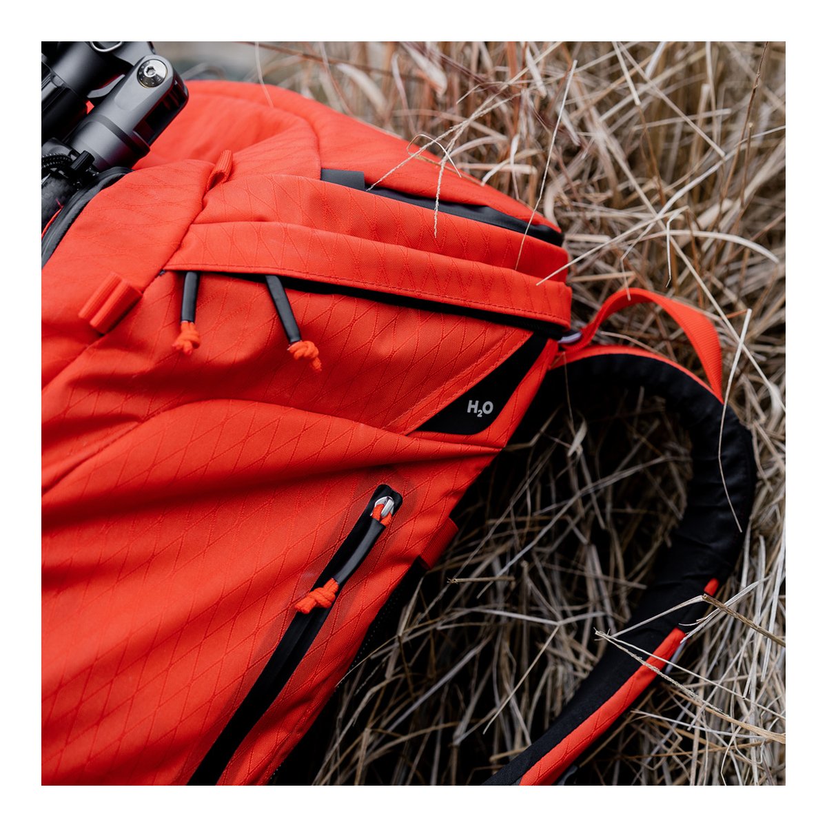 F-Stop DuraDiamond Tilopa - Magma - Detailansicht: Roter Outdoor-Rucksack mit H₂O-Fach, Reißverschlüssen und montiertem Stativ auf trockenem Gras. F-Stop DuraDiamond Tilopa - Magma - Detailansicht: Roter Outdoor-Rucksack mit H₂O-Fach, Reißverschlüssen und montiertem Stativ auf trockenem Gras.