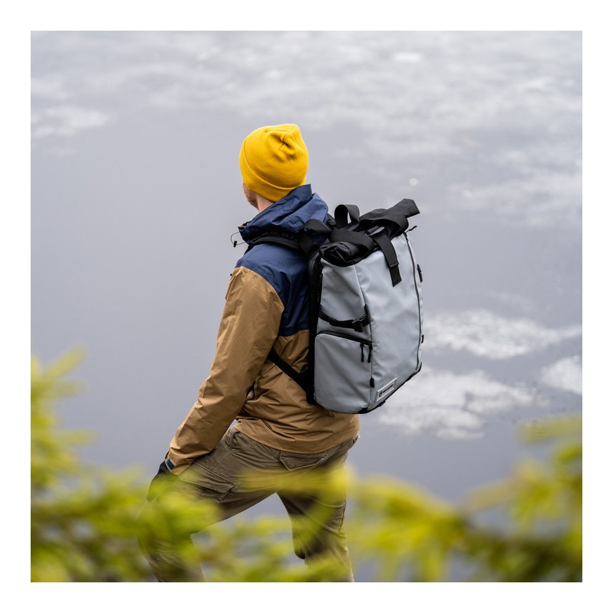 WANDRD PRVKE 41-Liter DisNet Grey V3 - Anwendungsbeispiel: Rückansicht eines Mannes mit grauem Roll-Top-Rucksack am Seeufer.