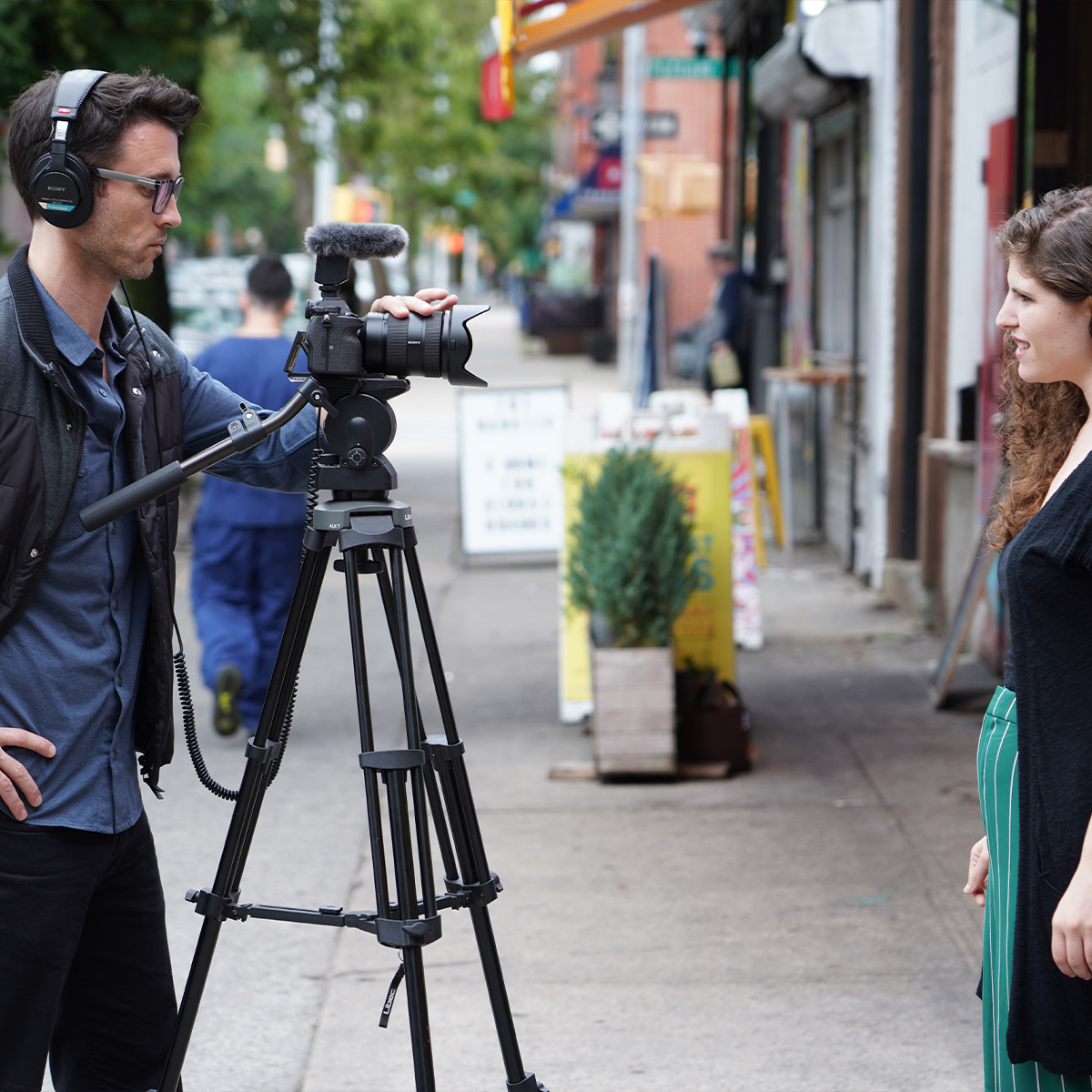 Mann filmt eine junge Frau mit  einer Sony Alpha Kamera an die das Sony ECM-B10 Mikrofon mit Windschutz angeschlossen ist auf einer Einkaufsstraße