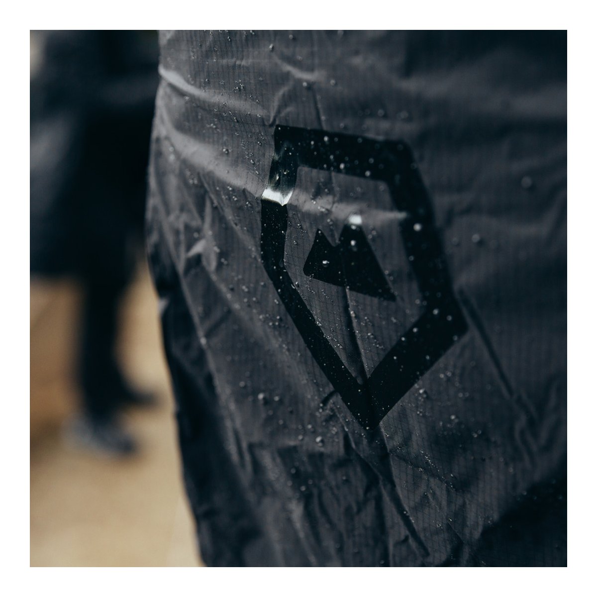 WANDRD RAINFLY - Detail: Wassertropfen auf schwarzem wasserdichtem Stoff mit Logo.