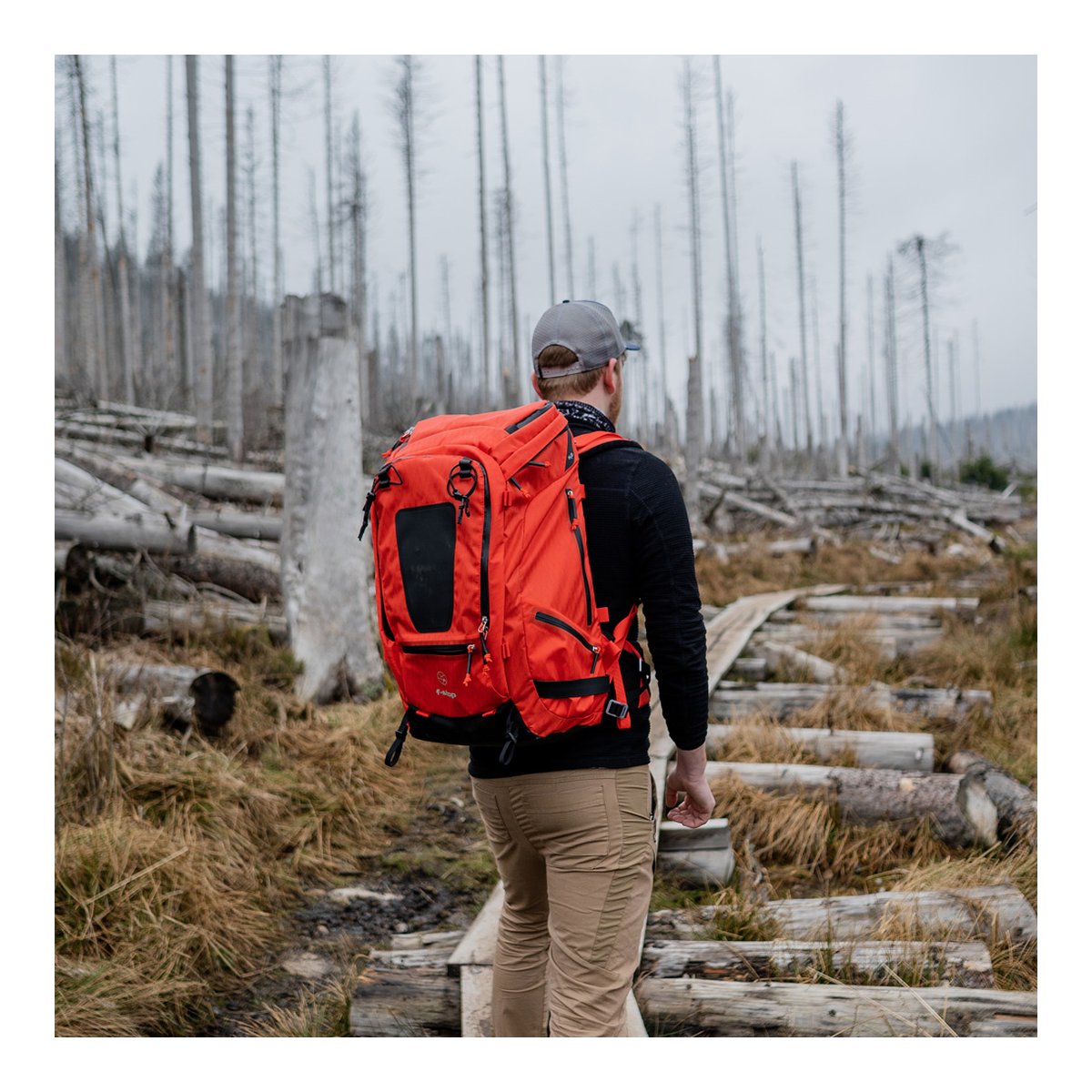 F-Stop DuraDiamond Tilopa - Magma - Anwendungsbeispiel: Rückansicht einer Person mit orangem Rucksack beim Wandern durch einen kargen Wald. F-Stop DuraDiamond Tilopa - Magma - Anwendungsbeispiel: Rückansicht einer Person mit orangem Rucksack beim Wandern durch einen kargen Wald.