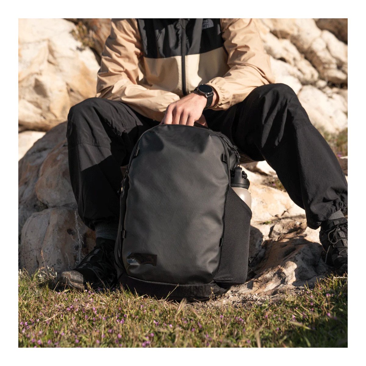 WANDRD NIMBUS 18 Black - Anwendungsbeispiel: Schwarzer Rucksack mit Trinkflasche in Seitentasche, Person sitzt auf Felsen im Freien