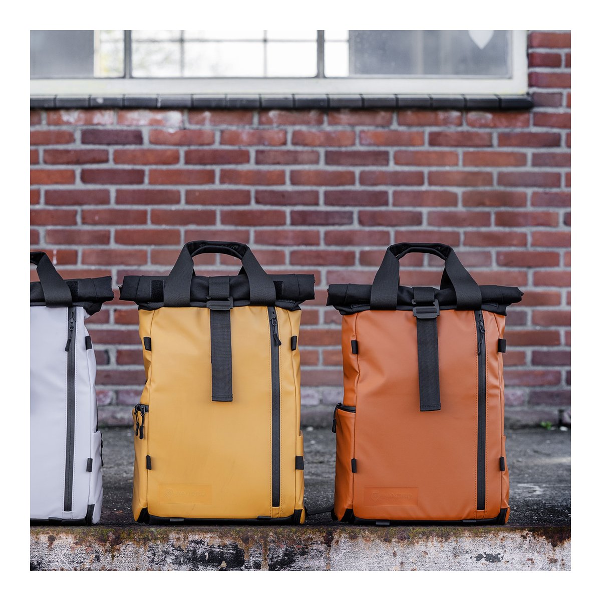 WANDRD PRVKE Lite 11-Liter Dallol Yellow - Frontansicht von drei Roll-Top-Rucksäcken in Weiß, Gelb und Orange vor einer Ziegelwand.