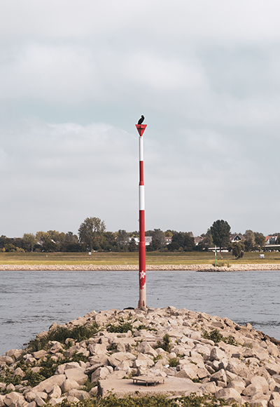 Minimalistische Fotografie – Vogel auf einer Wasserwegmarkierung am Fluss Eine Wasserwegmarkierung mit einem sitzenden Vogel an der Spitze, aufgenommen an einem Flussufer. Das Bild zeigt eine minimalistische Komposition mit natürlicher Tiefenwirkung.