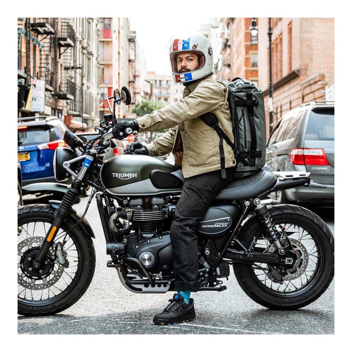 WANDRD HEXAD CARRYALL DUFFEL 40-Liter Wasatch Green - Anwendungsbeispiel: Mann mit olivgrünem Rucksack auf Motorrad in Stadtkulisse