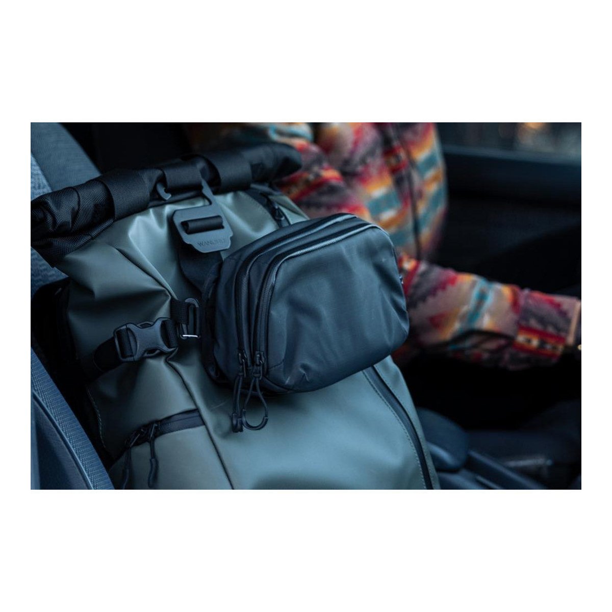 WANDRD Toiletry Bag Medium - {image_position}. Anwendungsbeispiel: Schwarze Zusatztasche befestigt an olivgrünem Rucksack im Auto.
