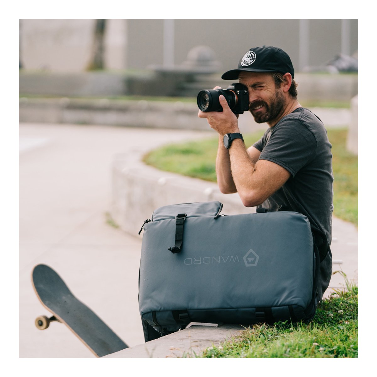 WANDRD HEXAD ACCESS DUFFEL Aegean Blue - Anwendungsbeispiel: Fotograf nutzt WANDRD Tasche im Freien, Skateboard daneben.