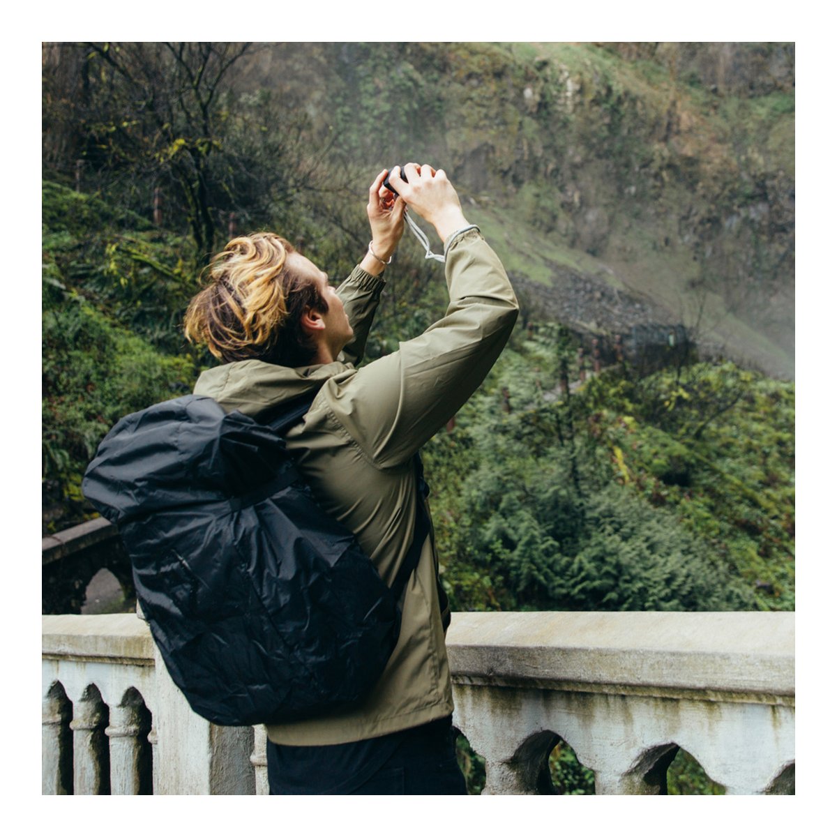 WANDRD RAINFLY - Anwendungsbeispiel: Person mit Rucksack-Regenschutz beim Fotografieren in der Natur.