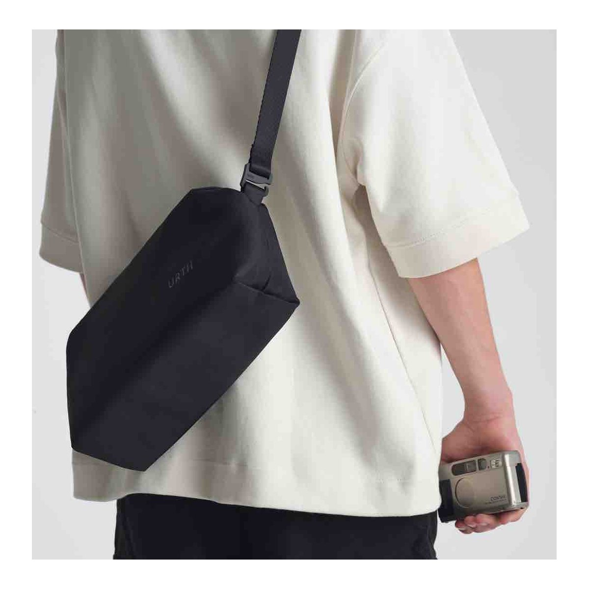 Urth Dolomite Camera Sling (Black) - Anwendungsbeispiel: Schwarze Umhängetasche am Rücken getragen, Person hält Kamera.