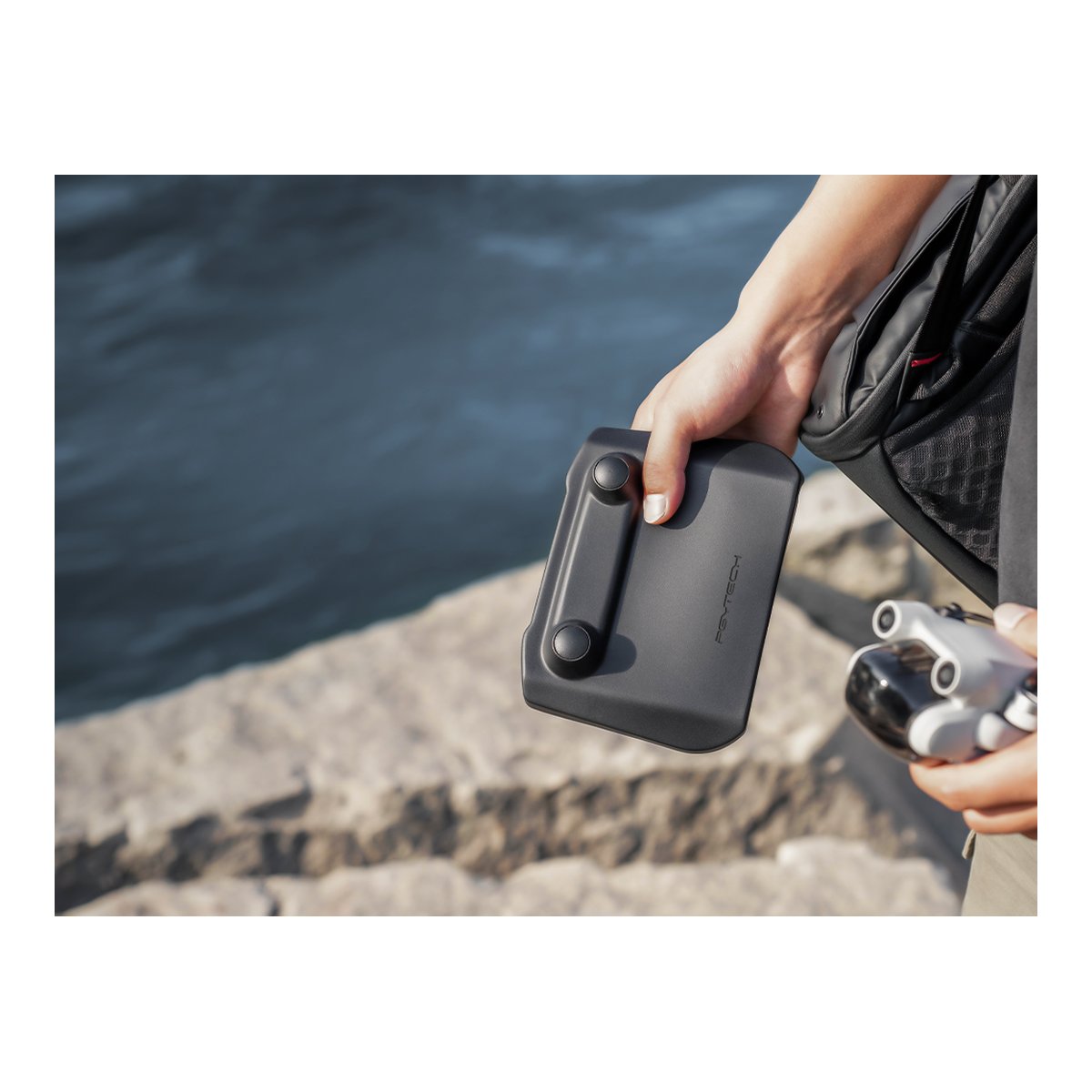 PGYTECH DJI RC Protector - Anwendungsbeispiel: PGYTECH Schutzzubehör mit Logo in der Hand eines Nutzers, Drohne daneben am Wasser.